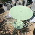 Salon de jardin en fer forgé 1950 : table en métal vert clair et chaises assorties sur un patio ensoleillé, entouré de seaux en métal et de plantes, évoquant le charme vintage. Brocante 93