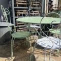 Salon de jardin en fer forgé 1950 : table et chaises en métal vert pâle posées sur une terrasse ensoleillée, avec des outils de jardin en arrière-plan. Brocante 93