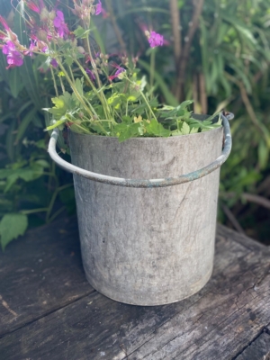 La Seau grande anse 1960, remplie de fleurs roses et de feuilles vertes, repose sur une surface en bois patinée à l'extérieur, capturant le charme vintage. Brocante 93