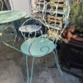 Le Salon de jardin en fer forgé 1960, doté d'une chaise en métal vert clair à volutes décoratives et d'une table assortie, apporte un charme vintage à votre terrasse en pierre. Brocante 93