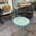 Une chaise de jardin en métal forgé bleu clair de style Salon de jardin des années 1960 avec un dossier décoratif se dresse sur un patio en pierre à côté de caisses en bois. Brocante 93