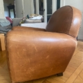 Une paire de fauteuils Club 1940 en cuir marron repose sur un parquet en bois, ajoutant un charme intemporel à un salon cosy. Brocante 93