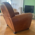 La paire de fauteuils Club 1940, en cuir marron, repose sur un parquet dans le salon avec une télévision, un miroir et un mur vert en arrière-plan. Brocante 93