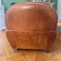 Une paire de fauteuils Club 1940 en cuir marron usé repose sur un plancher en bois, tourné vers l'extérieur, avec un mur vert et une cheminée en arrière-plan. Brocante 93