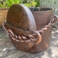 Un pot en métal rouillé avec un couvercle et une chaîne, posé sur une surface en bois usée par les intempéries, en plein air. Brocante 93