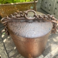 Un récipient en métal rouillé avec une anse en chaîne est posé sur une surface en bois usée par le temps, avec des plantes en arrière-plan. Brocante 93