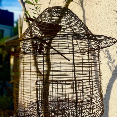 Une Cage déco jardin suspendue à l'extérieur contre un mur ensoleillé, avec une branche à l'intérieur, ce qui ajoute une charmante décoration extérieure à la scène. Brocante 93