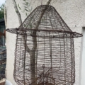 Une grande cage à oiseaux vide en fil de fer rouillé de Cage déco jard est suspendue à l'extérieur près d'un arbre et d'un mur texturé, apportant un style unique à votre jardin. Brocante 93