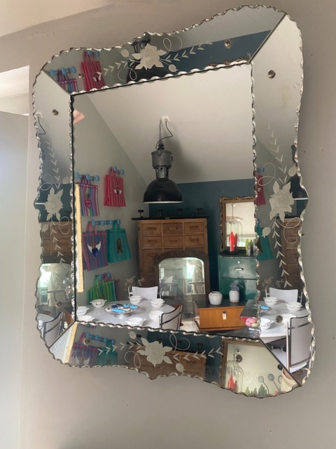 Le Miroir biseauté 1950 représente une salle à manger avec une table dressée et des serviettes colorées au mur, apportant un charme vintage au décor. Brocante 93