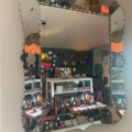 Un Miroir biseauté 1950 reflète l'intérieur d'une boutique confortable et éclectique remplie d'étagères, de lampes et de trésors uniques exposés. Brocante 93