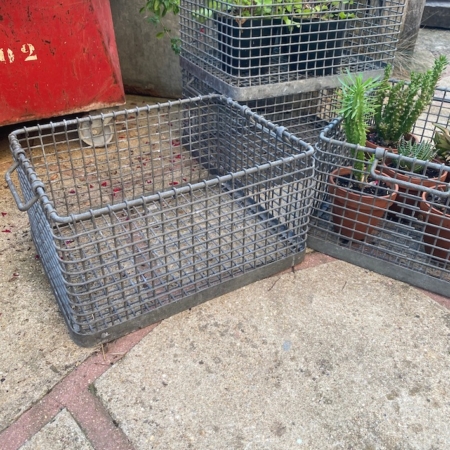 Un panier métallique des Caisses industrielles 1960 et des plantes en pot reposent sur un patio en béton ; le panier vide au premier plan ajoute un charme à la décoration vintage. Brocante 93