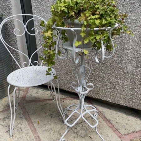 Chaise en fer forgé blanc et support de plantes assorti avec du lierre dans un pot, sur un patio en pierre près d'un mur texturé. Brocante 93