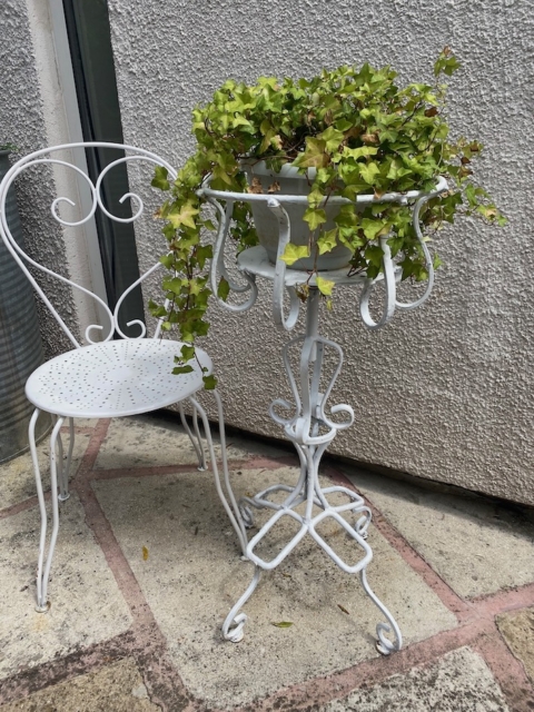 Chaise en fer forgé blanc et support de plantes assorti avec du lierre dans un pot, sur un patio en pierre près d'un mur texturé. Brocante 93