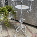Support de plantes en métal blanc sur un patio en pierre à côté d'une chaise blanche tenant une plante de lierre en pot. Brocante 93