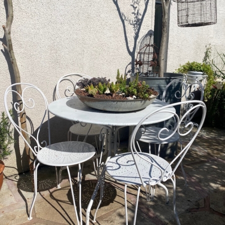 La table et les chaises de patio en métal blanc du Salon de jardin 1960 sont placées dans un coin de jardin ensoleillé, surmontées d'une jardinière de plantes grasses. Brocante 93