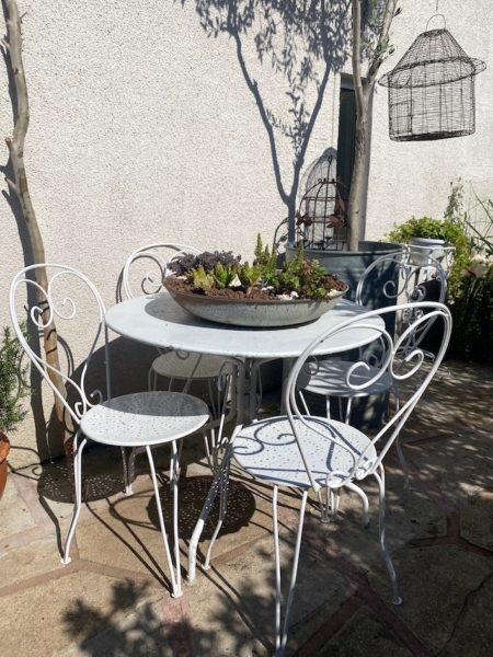 La table et les chaises de patio en métal blanc du Salon de jardin 1960 sont placées dans un coin de jardin ensoleillé, surmontées d'une jardinière de plantes grasses. Brocante 93