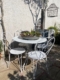 La table et les chaises de patio en métal blanc du Salon de jardin 1960 sont placées dans un coin de jardin ensoleillé, surmontées d'une jardinière de plantes grasses. Brocante 93