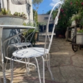 Le Salon de jardin 1960 - une chaise et une table en métal blanc disposées sur un patio en pierre ensoleillé, entouré d'une végétation luxuriante - confère un charme vintage à tout espace extérieur. Brocante 93