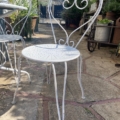 Le Salon de jardin 1960, une chaise en métal blanc avec une assise ronde et un dossier décoratif, est placé sur un patio en pierre au milieu d'un jardin luxuriant. Brocante 93