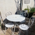 L'ensemble Salon de jardin 1960, composé d'une table et de trois chaises en métal blanc, est disposé sur un patio ensoleillé avec des plantes luxuriantes et une cage à oiseaux en fil de fer suspendue. Brocante 93