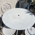 Le Salon de jardin 1960 est un ensemble table et chaises de patio en métal blanc au design perforé vintage des années 1960, placé à l'extérieur près d'une grande jardinière. Brocante 93