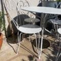 La table et les chaises bistro en métal blanc du Salon de jardin 1960 projettent des ombres élégantes sur un patio ensoleillé avec des plantes en pot, évoquant un charme classique - idéal pour tout salon de jardin ou mobilier extérieur. Brocante 93