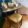 Une petite table en bois avec de la vaisselle et des vases en céramique est placée à côté d'un Tabouret paille vintage 1960 sur un sol en bois, créant un charmant look vintage. Brocante 93