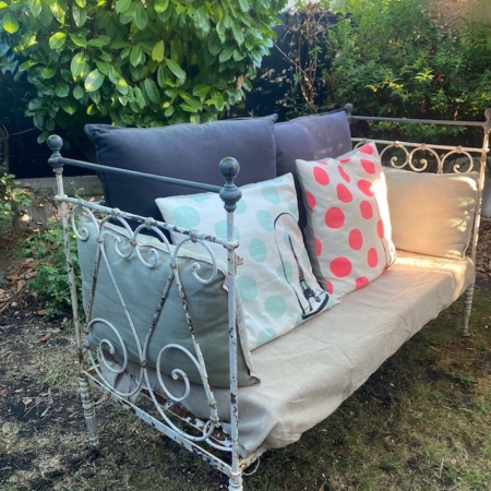 Un Lit en fer forgé 1950 vintage avec des coussins à pois colorés se trouve dans un jardin ensoleillé, entouré de buissons verts. Brocante 93