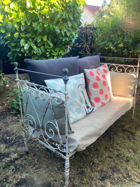 Un Lit en fer forgé 1950 vintage avec des coussins à pois colorés se trouve dans un jardin ensoleillé, entouré de buissons verts. Brocante 93