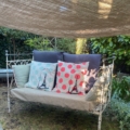 Le Lit en fer forgé 1950, installé en plein air avec des coussins de la Tour Eiffel sous un dais et la lumière du soleil, évoque le charme intemporel d'un jardin vintage. Brocante 93