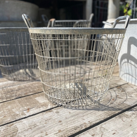 Un Panier métal grillagé 1950 repose sur une surface en bois usée par les intempéries à l'extérieur, la lumière du soleil projetant des ombres délicates. Brocante 93