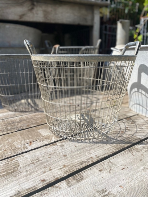 Un Panier métal grillagé 1950 repose sur une surface en bois usée par les intempéries à l'extérieur, la lumière du soleil projetant des ombres délicates. Brocante 93
