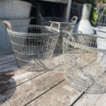 Deux paniers Panier métal grillagé 1950 sont posés à l'extérieur sur une surface en bois usée par les intempéries, avec des récipients en métal à l'arrière-plan. Brocante 93