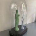 Le Globe de mariage 1950 présente deux petites statues, l'une verte et l'autre argentée, exposées sous un dôme de verre sur un socle noir contre un mur blanc. Brocante 93