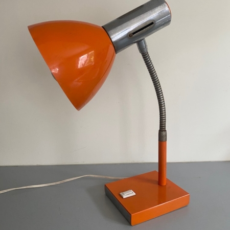 La Lampe articulée 1970, aux tons rétro orange et argent et à la base carrée, repose sur une surface grise contre un mur clair. Brocante 93