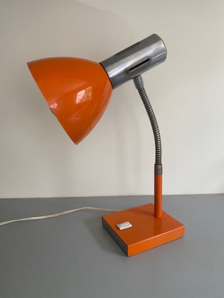 La Lampe articulée 1970, aux tons rétro orange et argent et à la base carrée, repose sur une surface grise contre un mur clair. Brocante 93