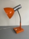La Lampe articulée 1970, aux tons rétro orange et argent et à la base carrée, repose sur une surface grise contre un mur clair. Brocante 93