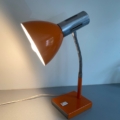 La Lampe articulée 1970 en orange et argent, avec un col flexible, est allumée et illumine une surface grise - un vrai clin d'œil au style vintage. Brocante 93