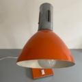 Une Lampe articulée 1970 en orange vintage avec une ampoule visible est exposée sur une surface grise contre un mur blanc. Brocante 93