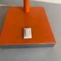 Gros plan de la base d'une Lampe articulée 1970 de couleur orange, comportant un interrupteur rectangulaire blanc, présenté sur une surface grise. Brocante 93