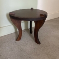 Le Repose pieds 1960 est un petit pouf à trois pieds en bois foncé avec un plateau rond, présenté sur un sol moquetté près d'un mur. Brocante 93