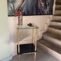 La Sellette verre et laiton 1950, avec ses pieds dorés, contient un vase avec des fleurs rouges à côté d'un escalier et d'une fresque murale de paons, reflétant le style chic des années 1950. Brocante 93