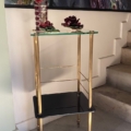 La Sellette verre et laiton 1950, une table d'appoint en verre et or avec des fleurs rouges décoratives, est placée près de l'escalier, à côté d'un tableau de paons. Brocante 93