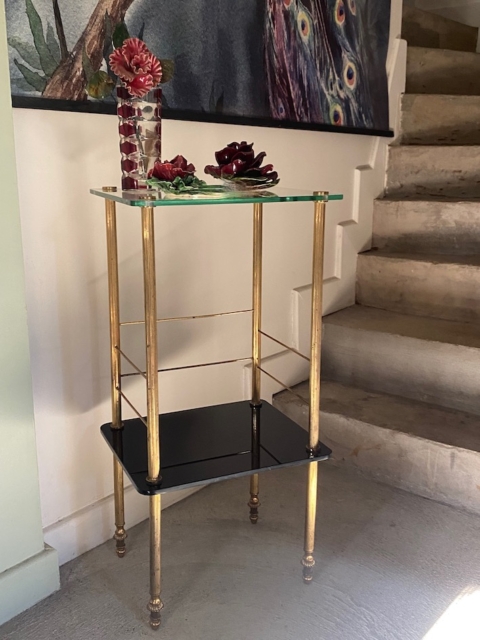 Une table d'appoint Sellette verre et laiton de 1950 avec des garnitures dorées et un décor floral rouge se trouve à côté d'un escalier en béton et d'un tableau de plumes de paon. Brocante 93
