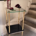 La Sellette verre et laiton 1950, une table d'appoint en verre or et noir ornée de fleurs rouges en céramique dans un vase, se trouve à côté d'un escalier en béton. Brocante 93