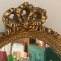 Gros plan sur le Miroir ovale 1950, un cadre décoratif ovale doré dans le style élégant des années 1950, avec une pièce légèrement floue se reflétant à l'arrière-plan. Brocante 93