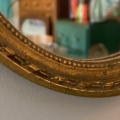 Gros plan sur le Miroir ovale 1950, un cadre ovale orné d'or reflétant une pièce floue à l'arrière-plan, évoquant l'élégance classique du milieu du siècle. Brocante 93