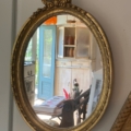 Le Miroir ovale 1950 présente un cadre doré et un style vintage, reflétant une armoire ancienne, une porte bleue ouverte et une partie d'une pièce intérieure. Brocante 93