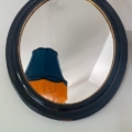 Un miroir mural noir ovale, le Miroir ovale noir 1950, reflète un abat-jour bleu à franges jaunes, évoquant le style classique des années 1950. Brocante 93