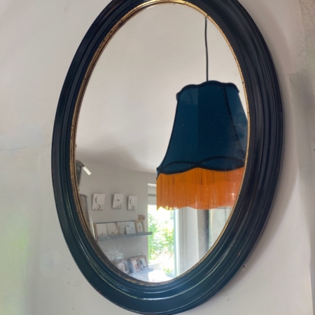 Le Miroir ovale noir 1950 reflète un abat-jour sombre avec une frange orange et fait partie d'une pièce lumineuse et confortable, évoquant l'élégance intemporelle des années 1950. Brocante 93